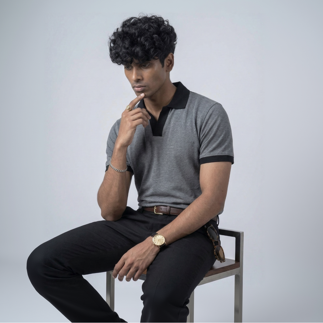 Man sitting on a chair wearing a gray polo shirt with black collar and black pants against a light gray background.